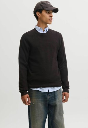 Homme portant un pull en maille noir par-dessus une chemise bleue boutonnée, un jean délavé et une casquette de baseball sombre, regardant sur le côté devant un fond uni.