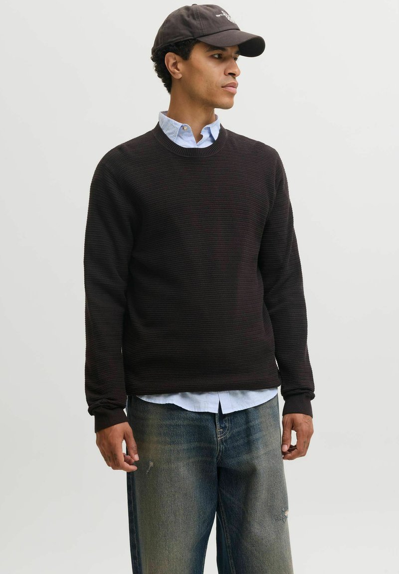 Homme portant un pull en maille noir par-dessus une chemise bleue boutonnée, un jean délavé et une casquette de baseball sombre, regardant sur le côté devant un fond uni.