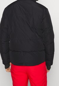 Svart pufferjacka med quiltade sektioner. Har en hög krage och ribbade cuffs. Bärs med röda byxor, vilket visar en snygg, lätt design.