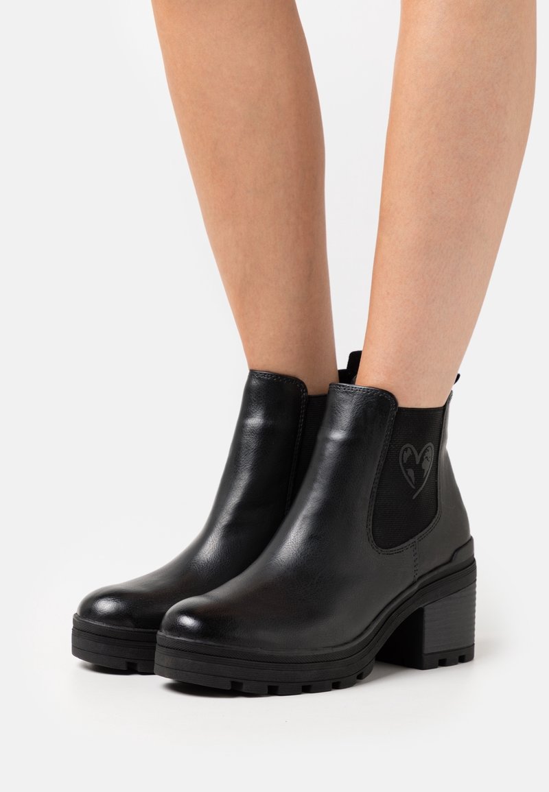Bottines en cuir noir avec talons blocs et détail cœur sur les panneaux latéraux élastiques, portées sur jambes nues sur fond blanc.