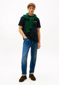Joven vestido con camiseta azul marina, jeans azules con dobles vueltas, zapatos marrones y un suéter verde sobre los hombros, contra un fondo blanco.