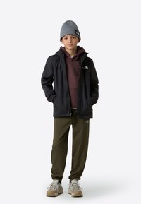 Schwarze wasserdichte Jacke über einem kastanienbraunen Hoodie, olivgrünen Hosen und grauer Strickmütze. Beige und graue Turnschuhe mit gemusterten Sohlen.