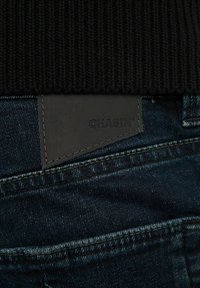 Svart ribbad tröja med mörkblå denimjeans, med en svart läderetikett med "CHASIN'" präglad. Närbild på tyg och sömdetaljer.