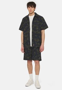 Set di camicia a maniche corte e pantaloni corti di colore blu scuro con motivi astratti verdi. T-shirt bianca semplice sotto e scarpe da ginnastica bianche indossate.