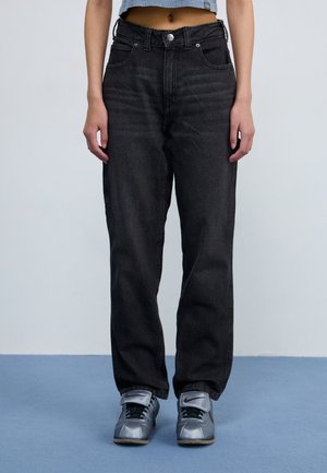 Person iført sorte jeans med høj talje, grå Nike-sneakers og en kort blå top, stående på en lyseblå gulv mod en hvid væg.