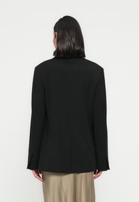 Άτομο με ίσια μαύρα μαλλιά, φοράει μαύρο σακάκι και μπεζ φούστα, γυρισμένο προς τα πίσω, σε απλό φωτεινό φόντο.