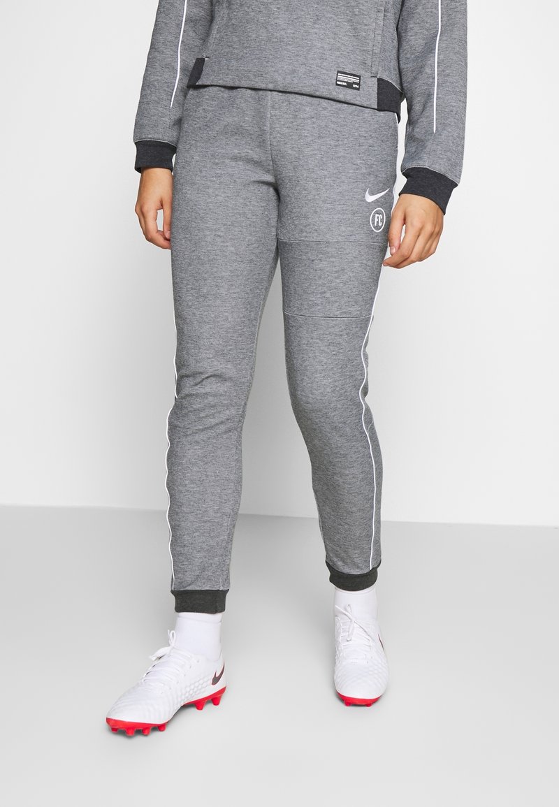 Personne portant un pantalon de survêtement Nike FC gris, des chaussettes blanches et des crampons blancs aux semelles rouges, debout sur un sol lisse gris clair.