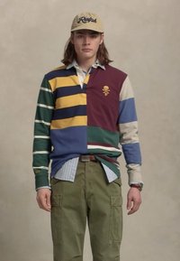 Kleurige gestreepte polo met lange mouwen in maroon, geel, blauw en groene secties. Olijfgroene cargobroek en een lichtblauwe gelaagde shirt eronder.