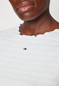 Nahaufnahme einer Person, die ein strukturiertes weißes Oberteil trägt, mit einem kleinen rechteckigen roten, weißen und marineblauen Logo, das mittig unter dem Ausschnitt platziert ist.
