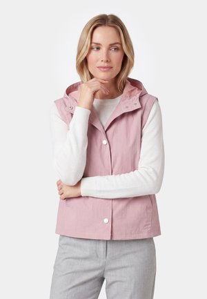 Kvinde med blondt hår iført en pink ærmeløs vest over en hvid langærmet skjorte og lysegrå bukser, poserer med hånden tæt på hagen.
