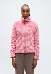 Γυναίκα που φοράει ροζ φλις μπουφάν με φερμουάρ και υφή, καθώς και καφέ παντελόνι, στέκεται μπροστά σε απλό ανοιχτό φόντο, κοιτάζοντας μπροστά.