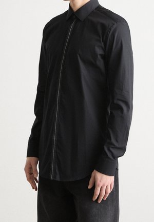 Man draagt een zwart shirt met lange mouwen en een verborgen rits aan de voorkant, gecombineerd met een zwarte broek, tegen een effen lichte achtergrond.