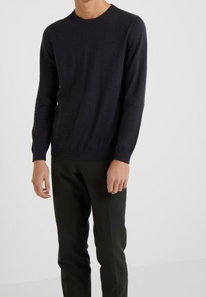 Uomo con maglione girocollo blu scuro e pantaloni neri in piedi davanti a uno sfondo chiaro e semplice.