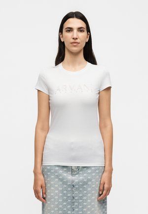 Młoda kobieta z długimi ciemnymi włosami w białym dopasowanym t-shircie Armani i jasnej jeansowej spódnicy z powtarzającym się białym wzorem logo „AX”.