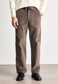 Pantaloni din catifea brown cu un design larg pe picior, având nervuri verticale proeminente, buzunare laterale și o curea neagră. Purtați cu pantofi negri.