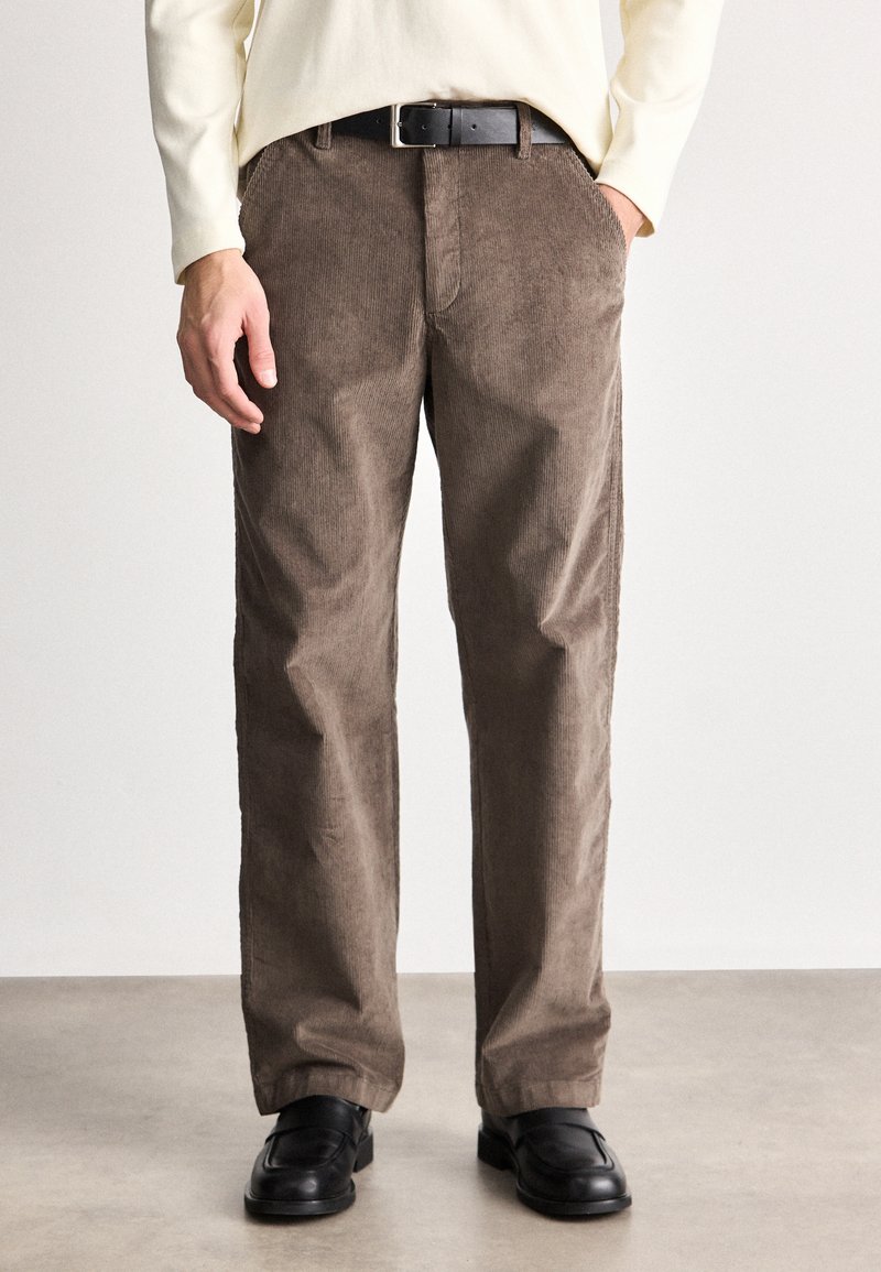 Pantaloni din catifea brown cu un design larg pe picior, având nervuri verticale proeminente, buzunare laterale și o curea neagră. Purtați cu pantofi negri.