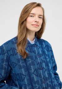 Niebieska kurtka pikowana o geometrycznym wzorze w ciemniejszym odcieniu niebieskiego, z okrągłymi guzikami i prążkowanym kołnierzem, wykonana z teksturowanej tkaniny.