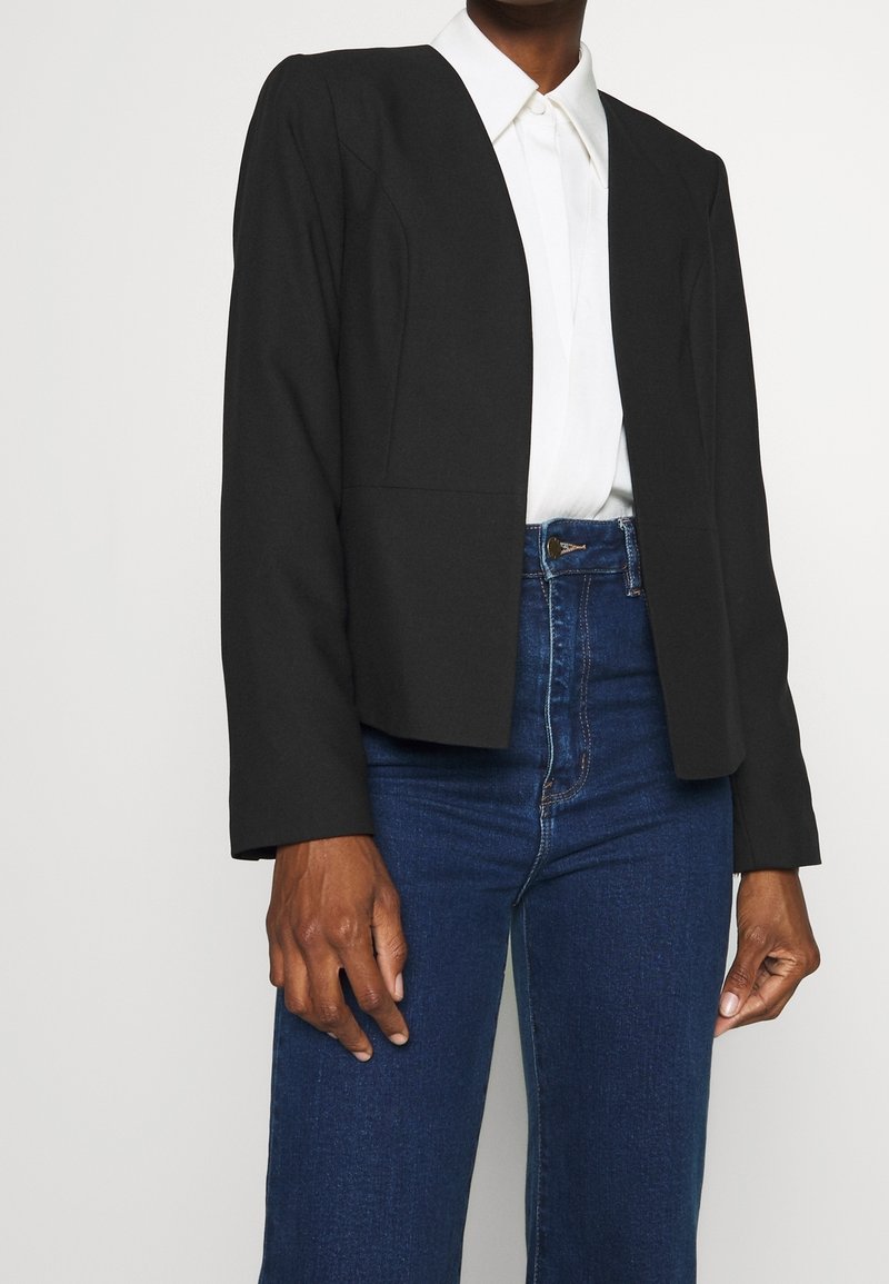Personne portant un blazer noir ajusté sur une chemise blanche à col, associé à un jean bleu taille haute devant un fond uni.