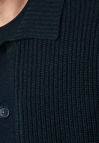 Pull en tricot noir texturé avec un design côtelé et un col. Dispose d'une fermeture à boutons et d'un tissu lisse au niveau du col et sur l'avant.