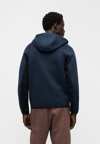 Tumši zils kapuču džemperis ar melnām sānu panelēm. Gluda tekstūra, piegulošs dizains un ribveida aproces. Neliels zīmola nosaukums pie apakšas.