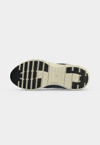 Semelle d'une basket avec grip en caoutchouc noir et beige, présentant un motif de bande de roulement segmenté et un logo embossé au centre.