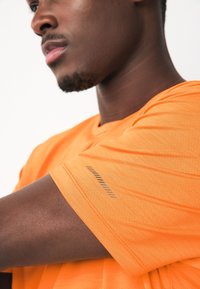 Maglietta sportiva arancione con tessuto strutturato, maniche corte e un motivo decorativo sulla manica. Vista ravvicinata del design.