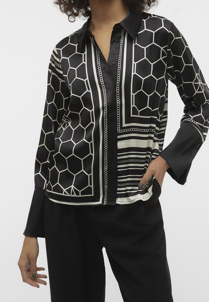 Personne portant une chemise boutonnée à motifs géométriques en noir et blanc à manches longues et un pantalon noir, main dans la poche sur fond blanc.