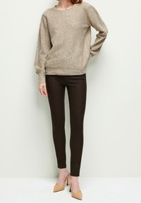 Beige gestrickter Pullover mit einem gesprenkelten Muster, kombiniert mit braunen, enganliegenden Leggings und beige hohen Mules. Glatte Textur und lockere Passform.