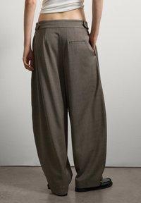 Persona vestida con pantalones anchos marrones, de pie con una mano en el bolsillo, visible desde la cintura hacia abajo sobre un fondo liso.