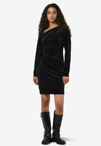 Svart fløyelskjole med tettsittende silhuett, ett-skulders design, lange ermer og prikket mønster; kombinert med knelang svarte støvler.