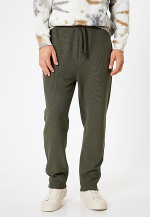 Olijf groene joggingbroek met een elastische tailleband en verstelbaar trekkoord. Textuurstof en taps toelopende benen. Draagt met witte sneakers.