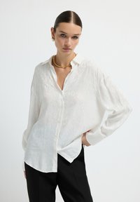 White, semi-sheer button-up shirt with leopard pattern, long sleeves, a collar, and a relaxed fit, paired with black trousers.