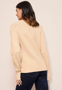 Bēšs pārāk liels džemperis ar ribotām apakšmalām un piedurkņu galiem, piedāvājot dekoratīvas pogas gar piedurknēm. Mīksta tekstūra, brīva piegrieza.