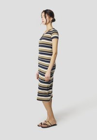 Sukienka midi w paski w kolorach granatowym, oliwkowym i beżowym; krótkie rękawy, dopasowana sylwetka, rozcięcie z boku, wykonana z miękkiego materiału; noszona z czarnymi, płaskimi sandałami.