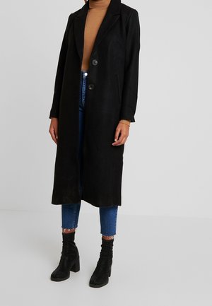 Personne portant un long manteau noir par-dessus un col roulé beige, un jean bleu et des bottines noires à talons, debout sur un fond gris uni.