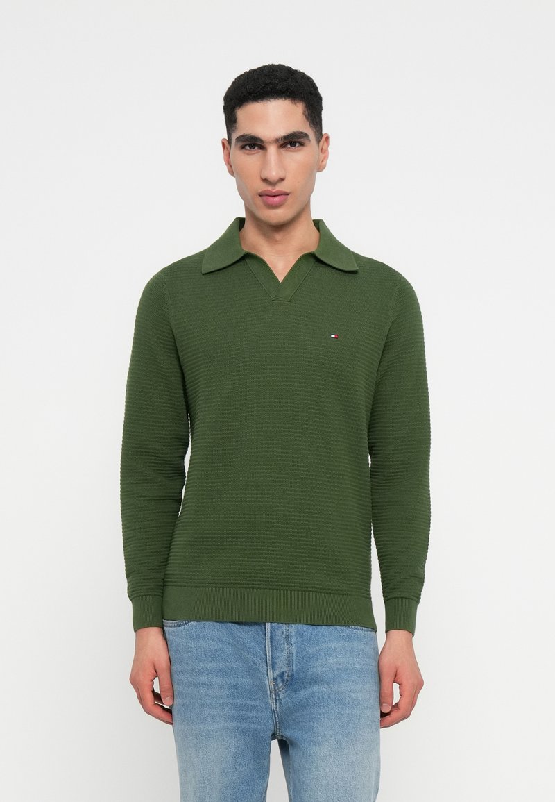 Zöld kötött pulóver gallérral és hosszú ujjal, textúrázott dizájnnal és egy kis logóval a mellkason. Kék farmernadrággal viselve.