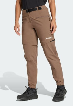 Pantalon de randonnée marron avec jambes zippées, chevilles élastiques, ceinture et poches avant, porté avec des chaussures de trail noires sur une personne debout.