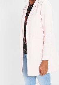 Light pink blazer with a structured design, featuring a single button closure and two front pockets, worn over a black floral top.