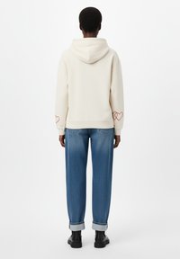 Kremveida kapuce ar sarkanu sirdīšu izšuvumu uz piedurknēm, kopā ar brīviem zila denimauga džinsiem, kuriem ir izlocīti gali un gluda tekstūra.