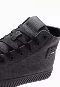 Zapatilla alta de lona negra con un acabado texturizado, cordones planos, suela de goma y un acento de logo negro en el lado.
