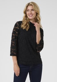 Top in pizzo nero con maniche a tre quarti, caratterizzato da un motivo floreale e un collo alto, indossato con jeans in denim scuri.