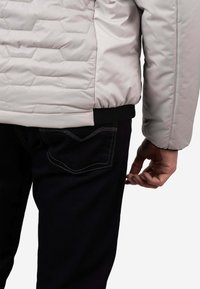 Chaqueta acolchada gris claro con patrón de acolchado hexagonal y dobladillo acanalado negro, combinada con jeans oscuros que presentan costuras de contraste y un bolsillo trasero.