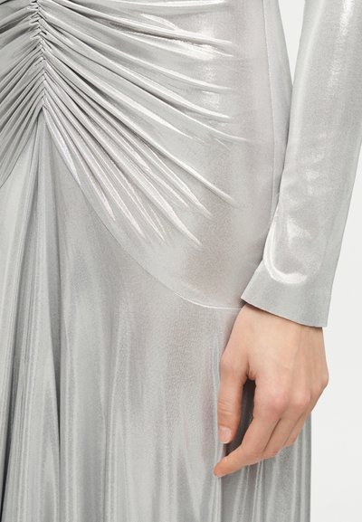 Κοντινό πλάνο ασημένιου μεταλλικού φορέματος με μακριά μανίκια, με σούρες και ρυτίδες, και ένα χέρι που ακουμπά απαλά στο πλάι.