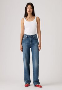 Νεαρή γυναίκα με μακριά σκουρόχρωμα μαλλιά, φορώντας λευκό τοπ χωρίς μανίκια, μπλε τζιν ψηλόμεσο και κόκκινα πλατφόρμες, στέκεται σε απλό φόντο.