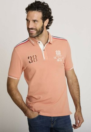 Homme aux cheveux bouclés portant un polo saumon avec le numéro 38 et un texte blanc, mains dans les poches, souriant et regardant à gauche.
