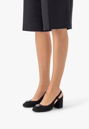 Nogi w czarnych sandałach na obcasie typu blok z ozdobnymi, okrągłymi guzikami, w połączeniu z czarną spódnicą do kolan.