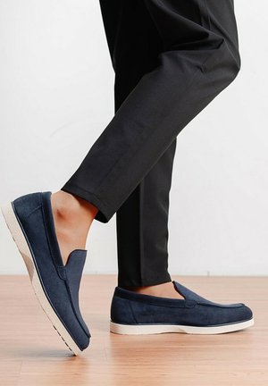 Personne portant des chaussures sans lacets bleu marine avec des semelles blanches, debout sur un sol en bois clair, un pied légèrement levé, vêtue d'un pantalon noir.