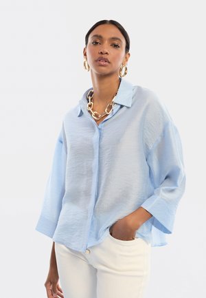 Donna che indossa una camicia oversize azzurro chiaro con bottoni, pantaloni bianchi, grandi orecchini a cerchio dorati e una collana a catena dorata spessa, mano in tasca.
