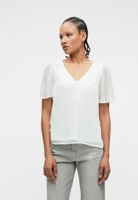 Λευκή μπλούζα με κοντά, διαφανή μανίκια και V-λαιμόκοψη. Το ύφασμα φαίνεται ελαφρύ και ημιδιαφανές, με ομαλή επιφάνεια.