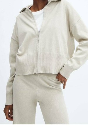 Žena v béžovém svetru na zip a odpovídajících vysokým kalhotám, s jednou rukou opřenou o bok na jednobarevném pozadí.